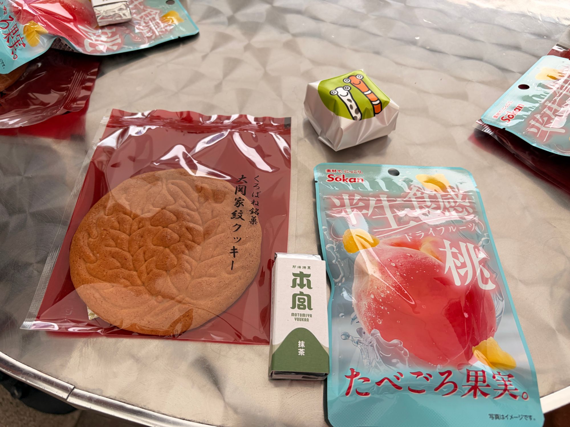 テーブルに並んだ家紋クッキー、抹茶の羊羹、桃のドライフルーツの袋